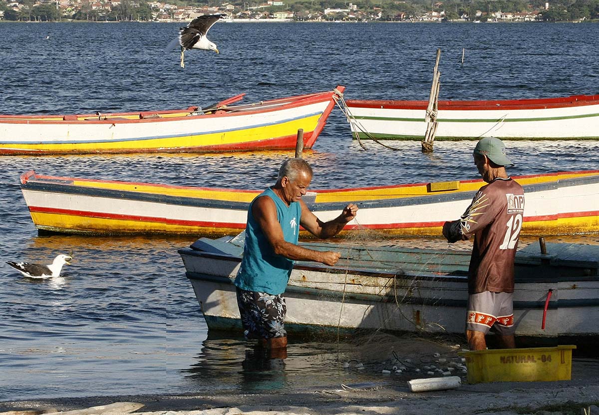 Dois homens dentro do mar pescam; ao fundo aparecem barcos