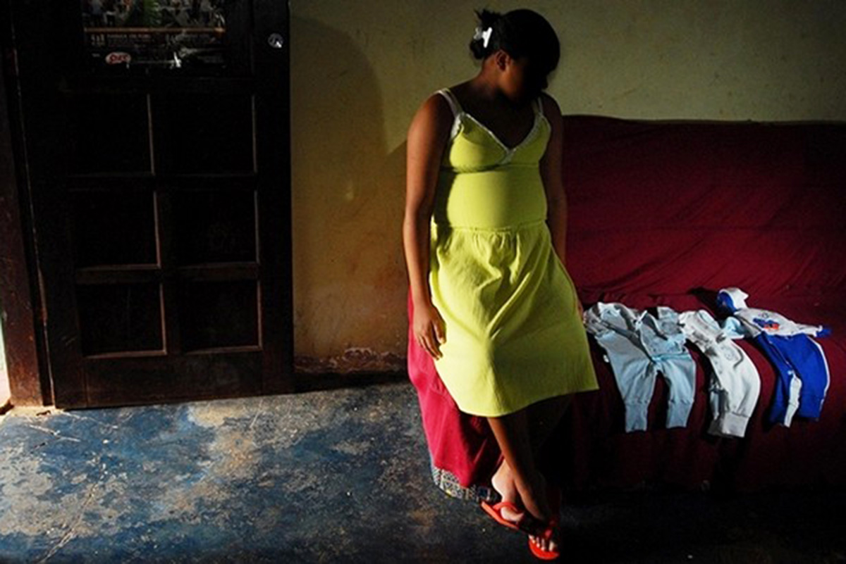 Jovem grávida usando vestido aparece encostada em sofá enquanto observa roupas de bebê 