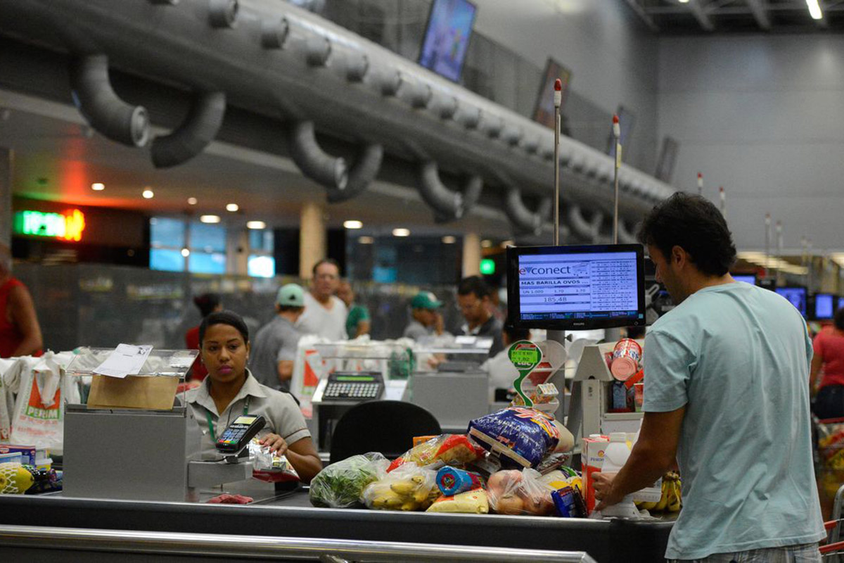 No caixa de supermercado, mulher sentada e homem em pé, e na esteira compras