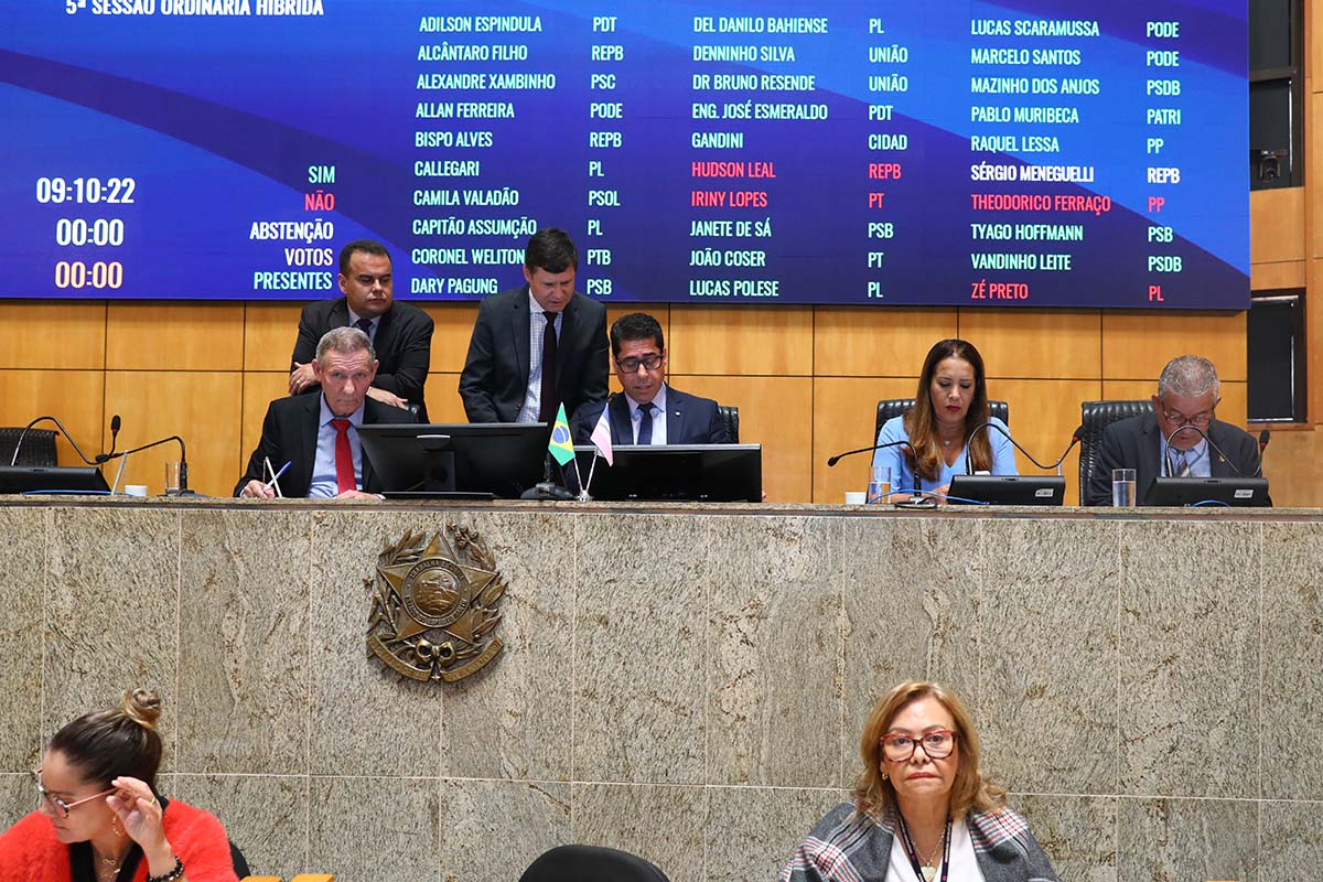 Pessoas em pé e sentadas atrás da mesa do Plenário e acima delas painel eletrônico azul