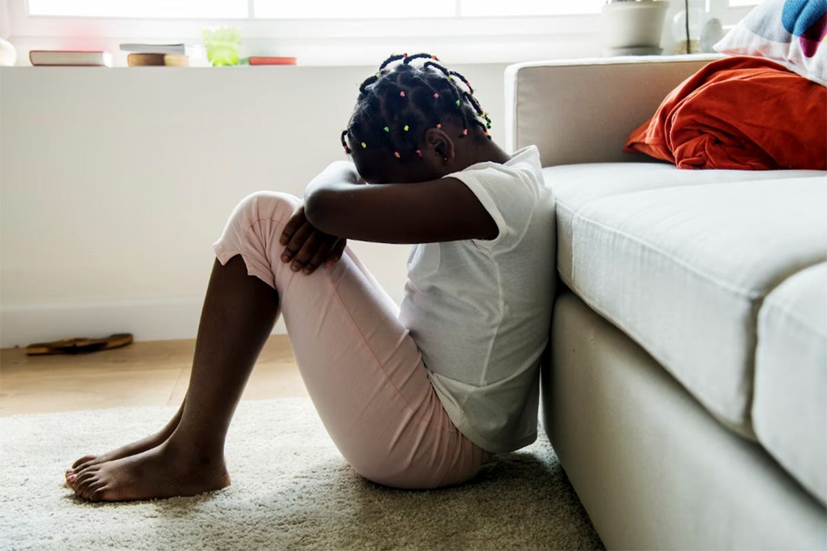Criança negra sentada apoiada em sofá com cabeça baixa abraça os joelhos
