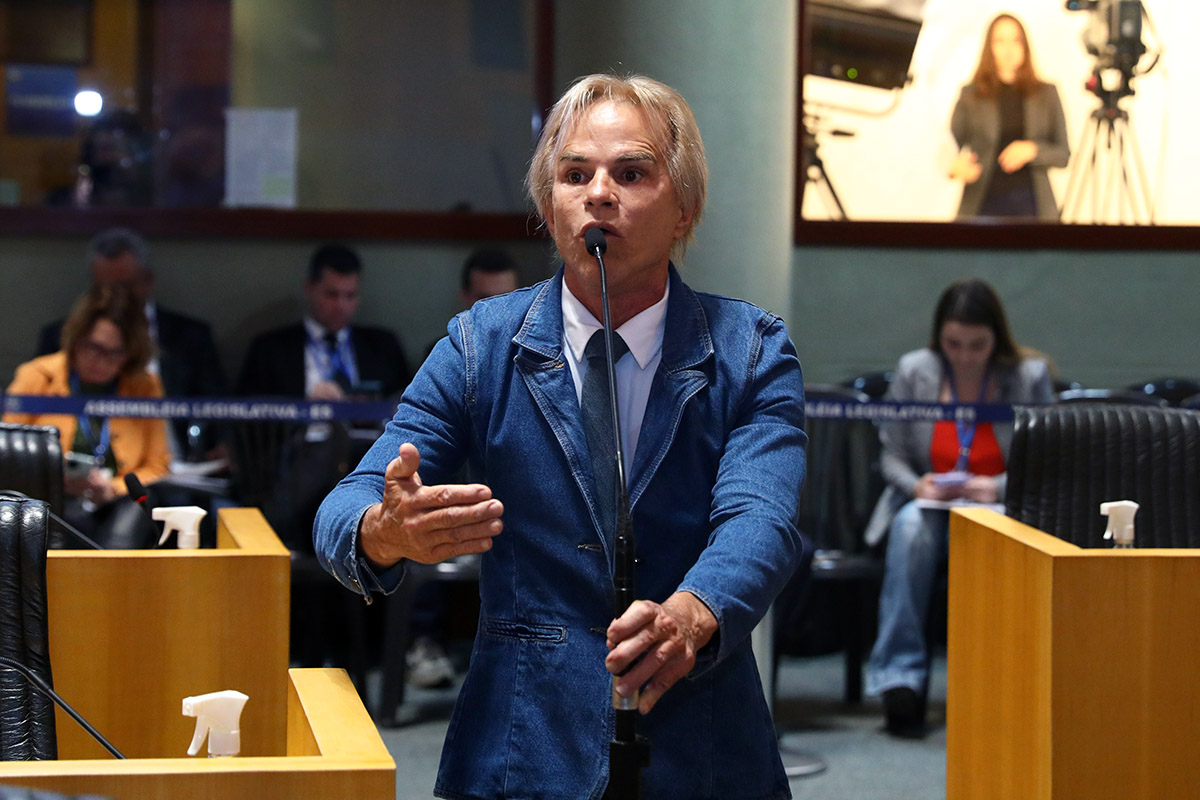 Homem de terno jeans, em pé ao microfone no Plenário da Assembleia